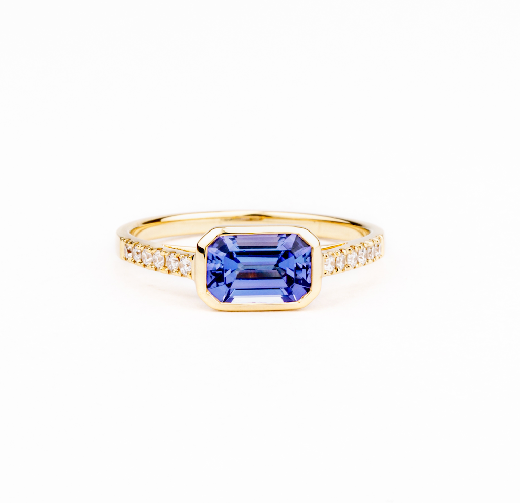 Satin 18k yellow gold ring with east-west emerald-cut tanzanite in a bezel setting and diamond-accented band