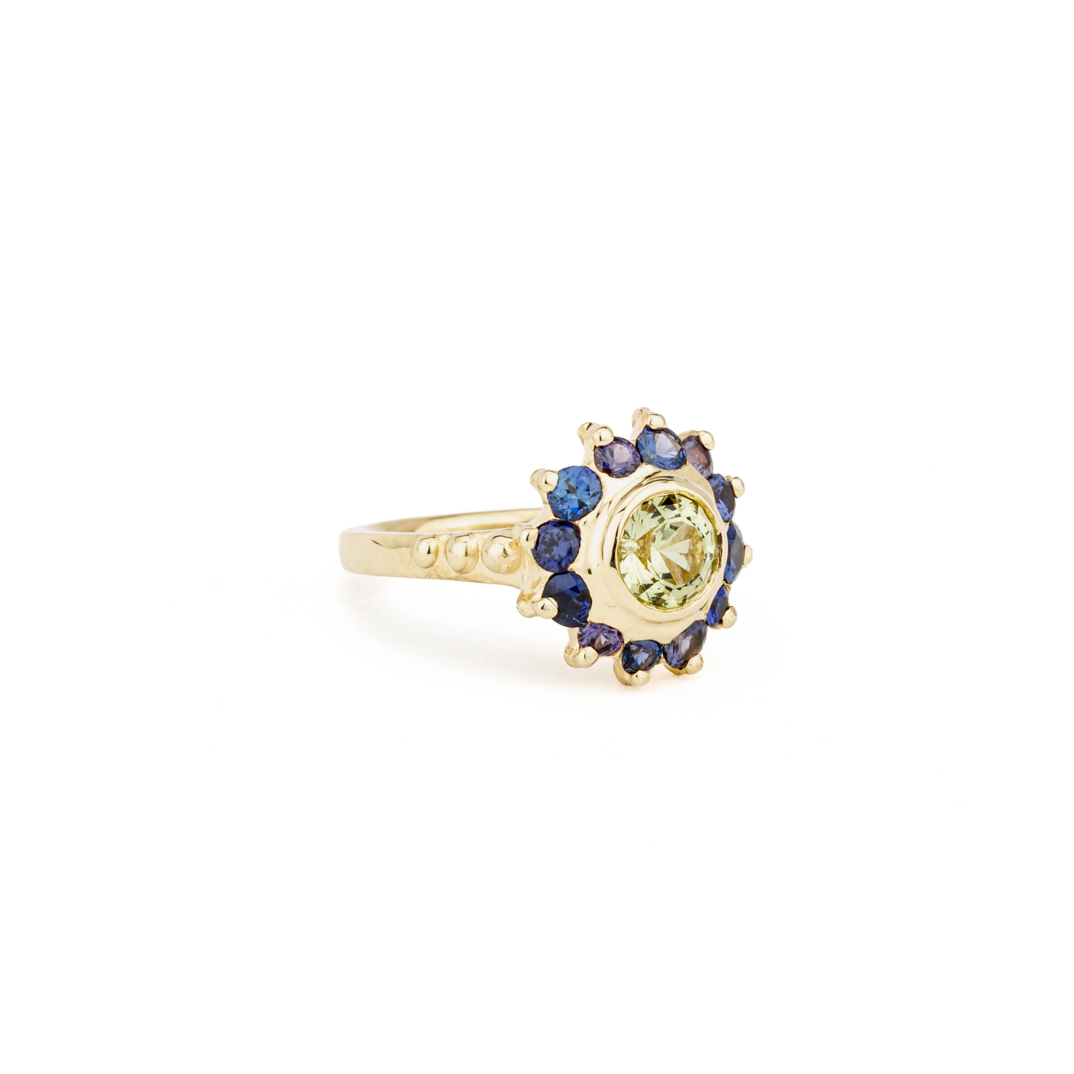 Yellow gold ring with a bezel-set green chrysoberyl center surrounded by a sunburst halo of blue and purple round sapphires on a beaded band.
