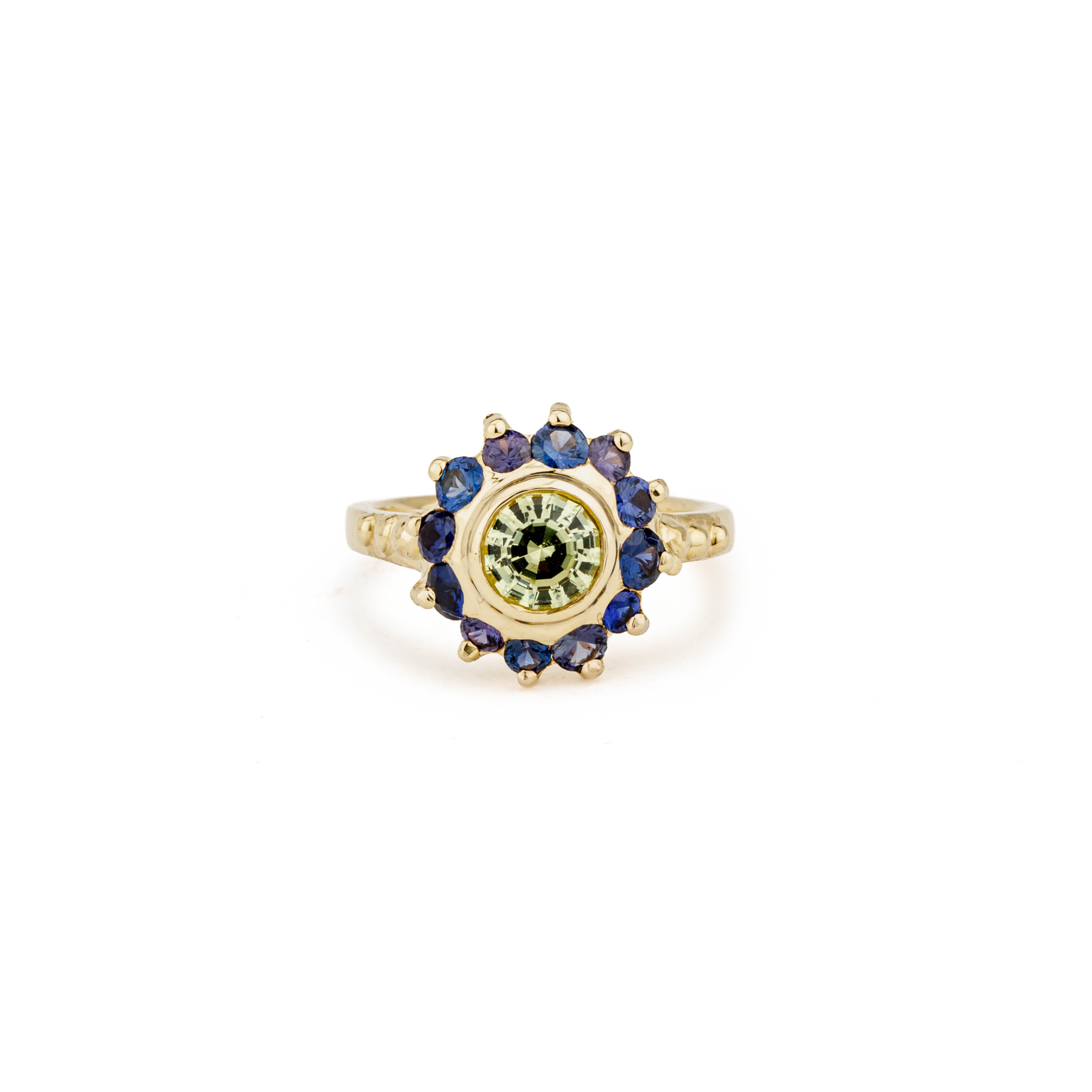 Yellow gold ring with a bezel-set green chrysoberyl center surrounded by a sunburst halo of blue and purple round sapphires on a beaded band.