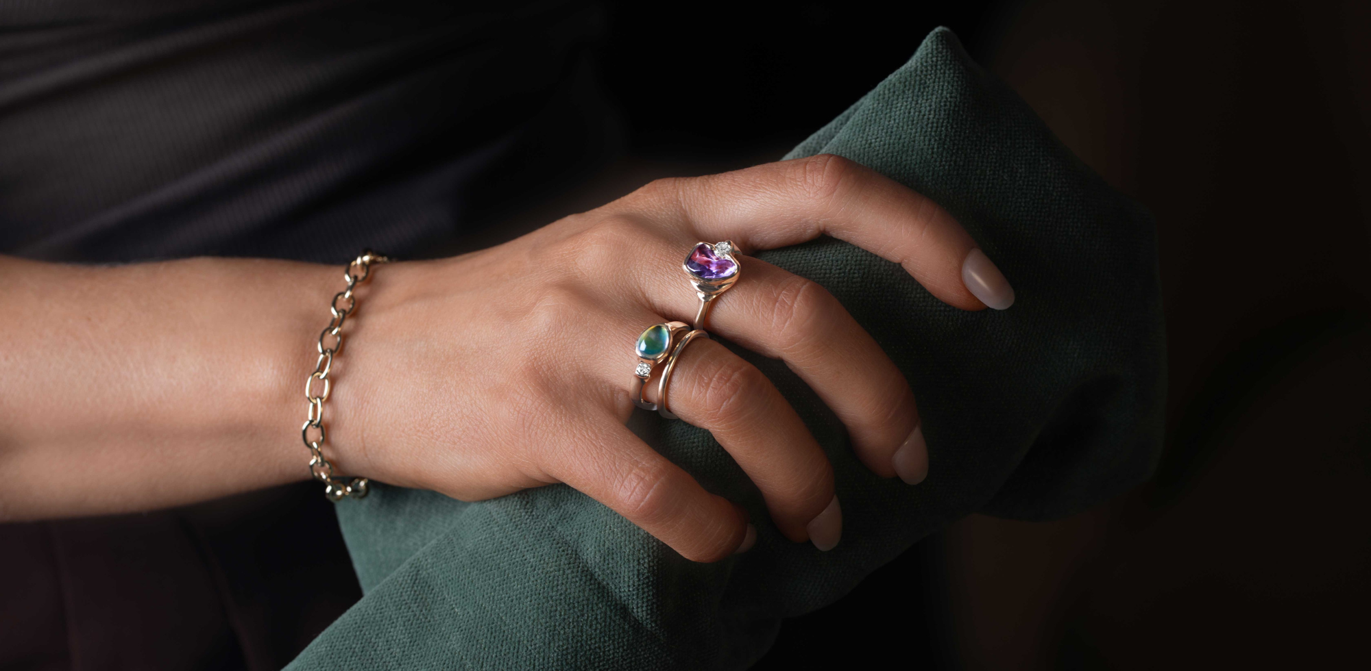 Close-up of a hand wearing multiple rings designed by deJonghe Jewelry with a moonstone and purple sapphire in 14k yellow gold on a dark background