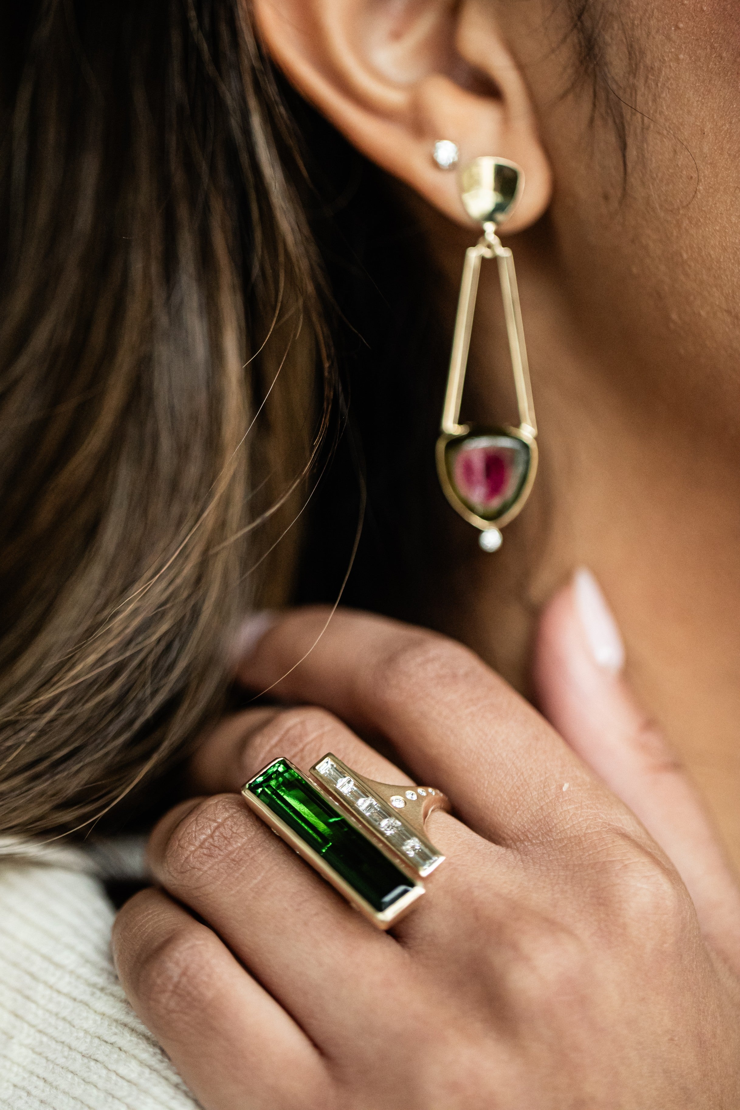 Close-up of a person wearing gold watermelon tourmaline earrings and a ring with a green tourmaline by deJonghe Original Jewelry.