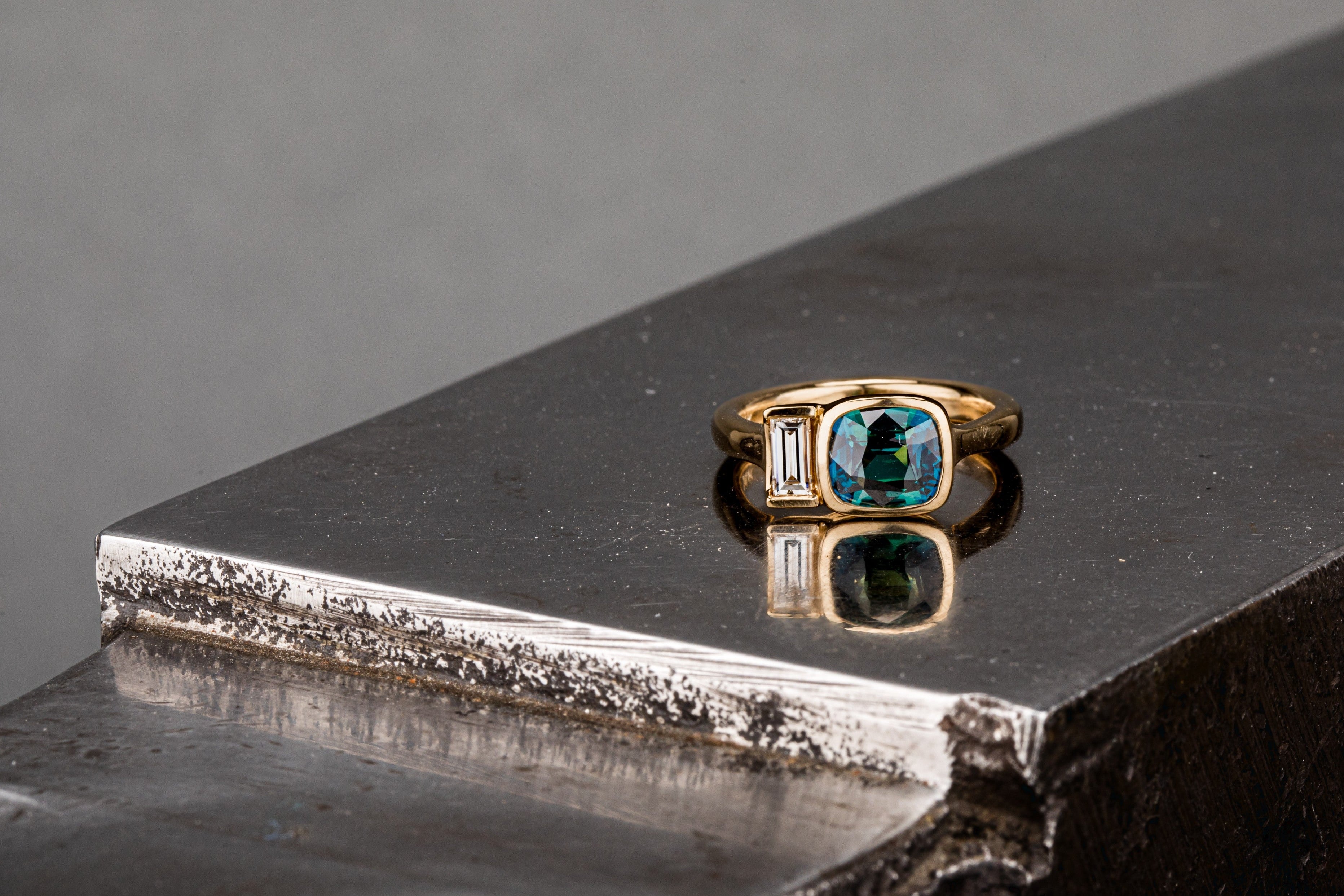 14k gold ring with a teal sapphire and baguette diamond photographed on an anvil